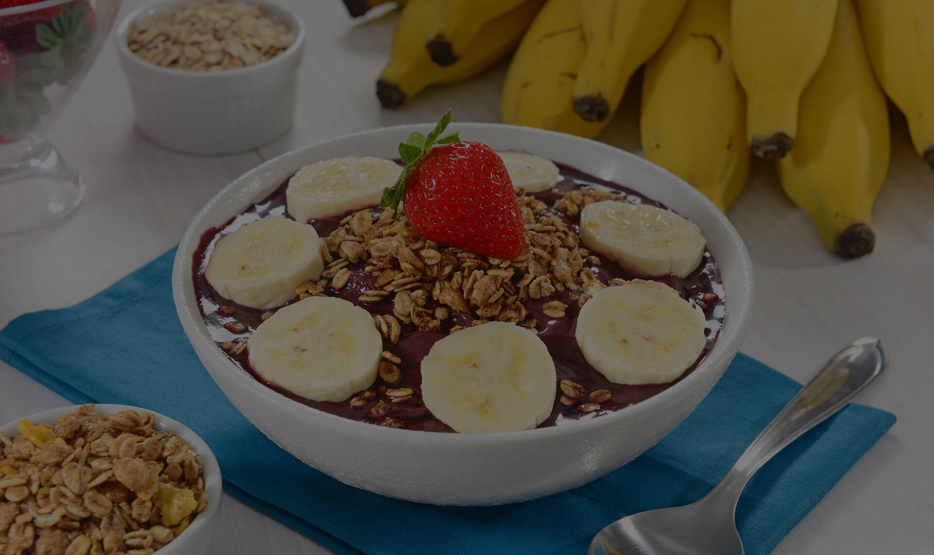 Açaì in a white bowl topped with oats, banana and strawberry with more fruit and oats in the background.